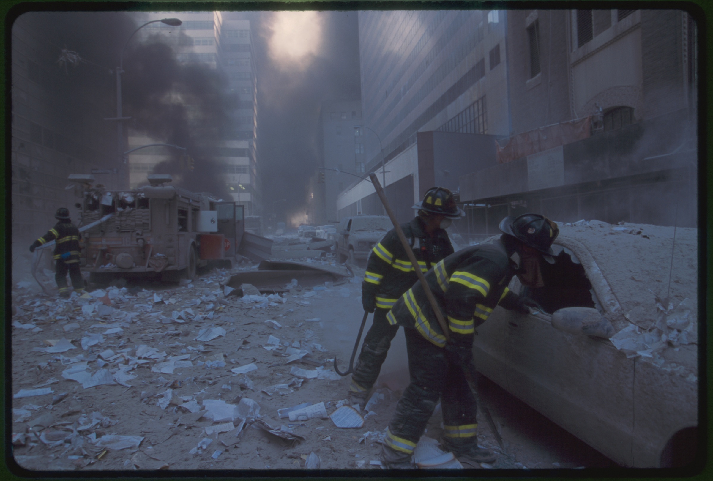 Two fire fighters looking into car while another New York City fire fighter pulls water hose from fire truck amid smoke and debris following September 11th terrorist attack on World Trade Center, New York City